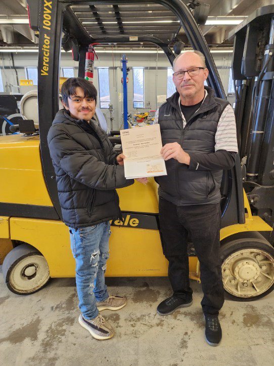 Rodmam, a 20-year-old, shakes the hand of Mr. Terrance, a middle-aged person, while holding a forklift certification certificate. They are standing in front of a yellow forklift.