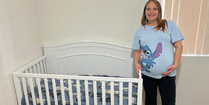 Pregnant woman in a light blue graphic tee standing next to a white crib.